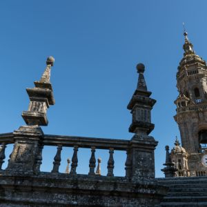 Crestería barroca de piedra en las cubiertas de la Catedral de Santiago, con la Torre del Reloj elevándose al fondo bajo un cielo azul despejado.