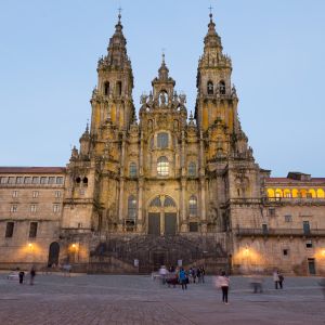 Fachada del Obradoiro iluminada al anochecer desde la plaza. Vista frontal de las torres barrocas y el gran espejo de vidrio central.
