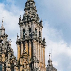 Detalle barroco de la Torre de las Campanas en la Catedral de Santiago. Estructura de piedra con campanas visibles y pináculos bajo un cielo azul.