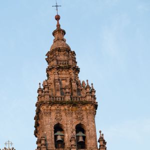 Detalle del remate superior de la Torre de las Campanas, mostrando la aguja de piedra y la cruz de bronce frente a un cielo azul despejado.