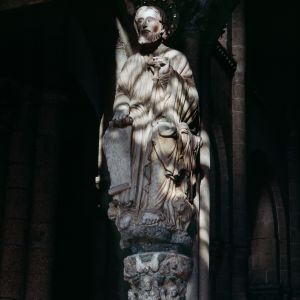 Santiago Apóstol en el parteluz del Pórtico de la Gloria iluminado por el atardecer, con báculo en tau y capitel de la Trinidad.